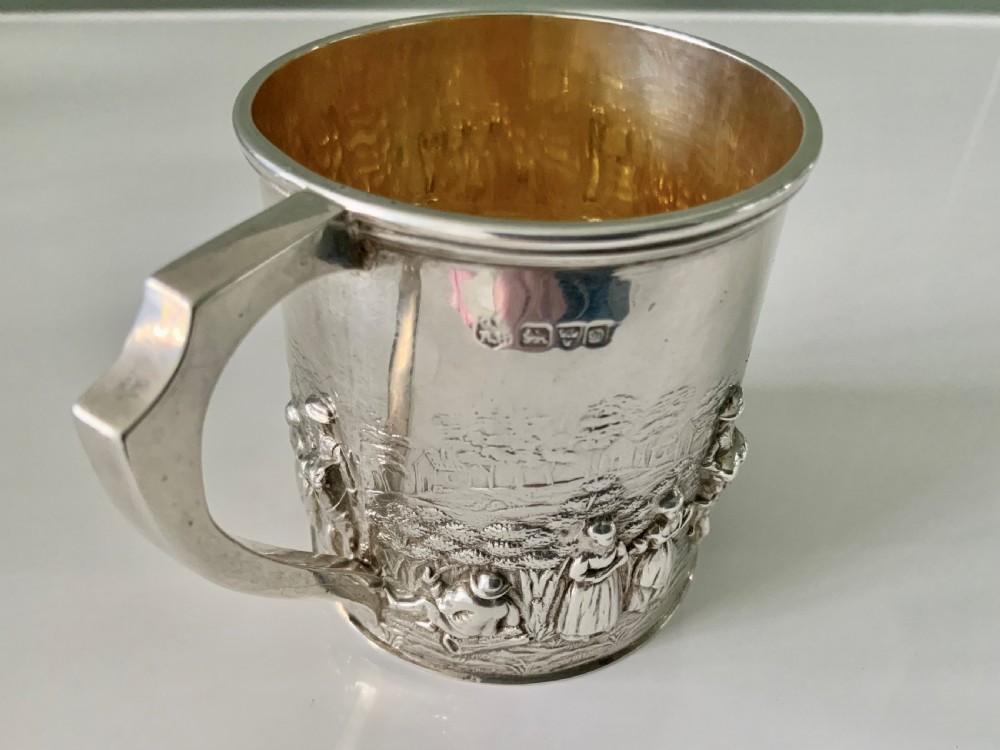 A small sterling silver christening mug decorated with embossed pastoral figures and a church landscape, featuring an angular side handle. View IV