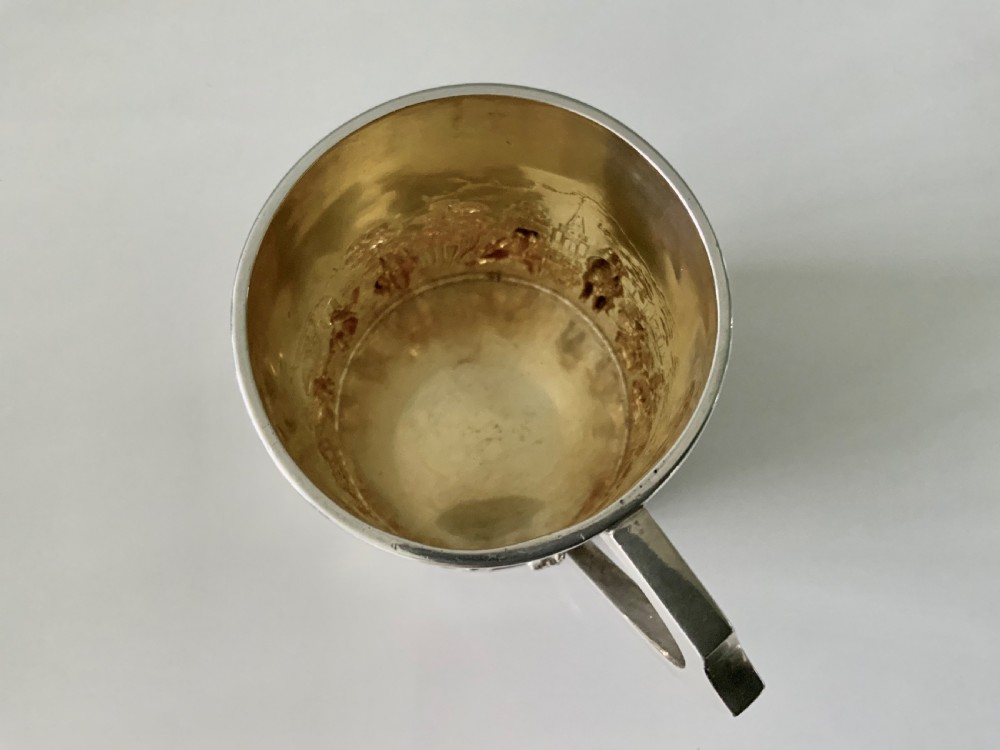 A small sterling silver christening mug decorated with embossed pastoral figures and a church landscape, featuring an angular side handle. View V