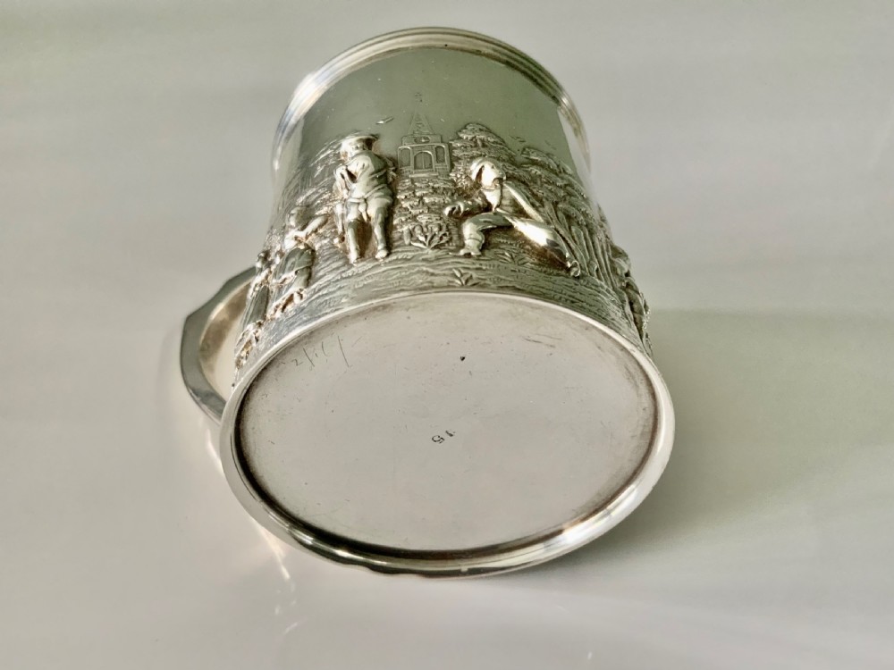 A small sterling silver christening mug decorated with embossed pastoral figures and a church landscape, featuring an angular side handle. View VI
