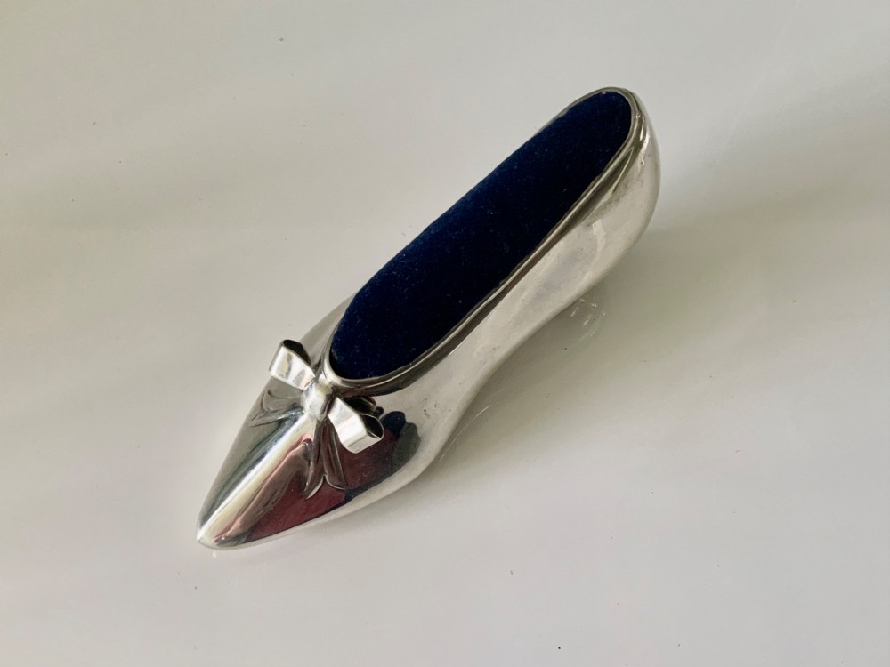 A silver novelty pin cushion shaped like a pointed shoe featuring a silver bow motif on the vamp and a navy blue velvet interior insert.