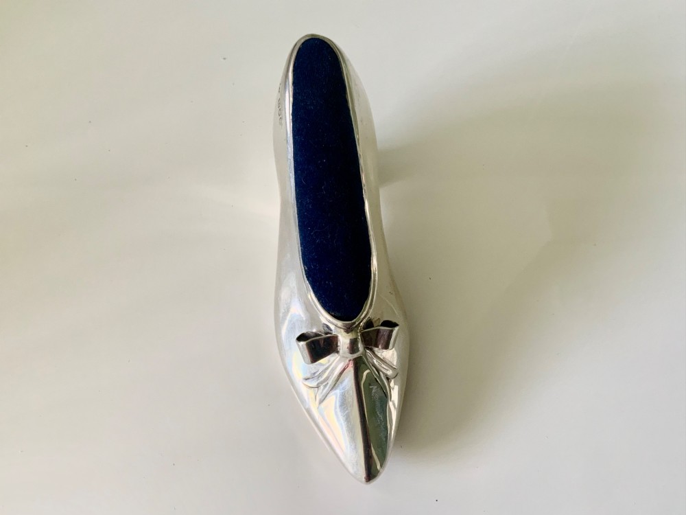 A silver novelty pin cushion shaped like a pointed shoe featuring a silver bow motif on the vamp and a navy blue velvet interior insert. View VI