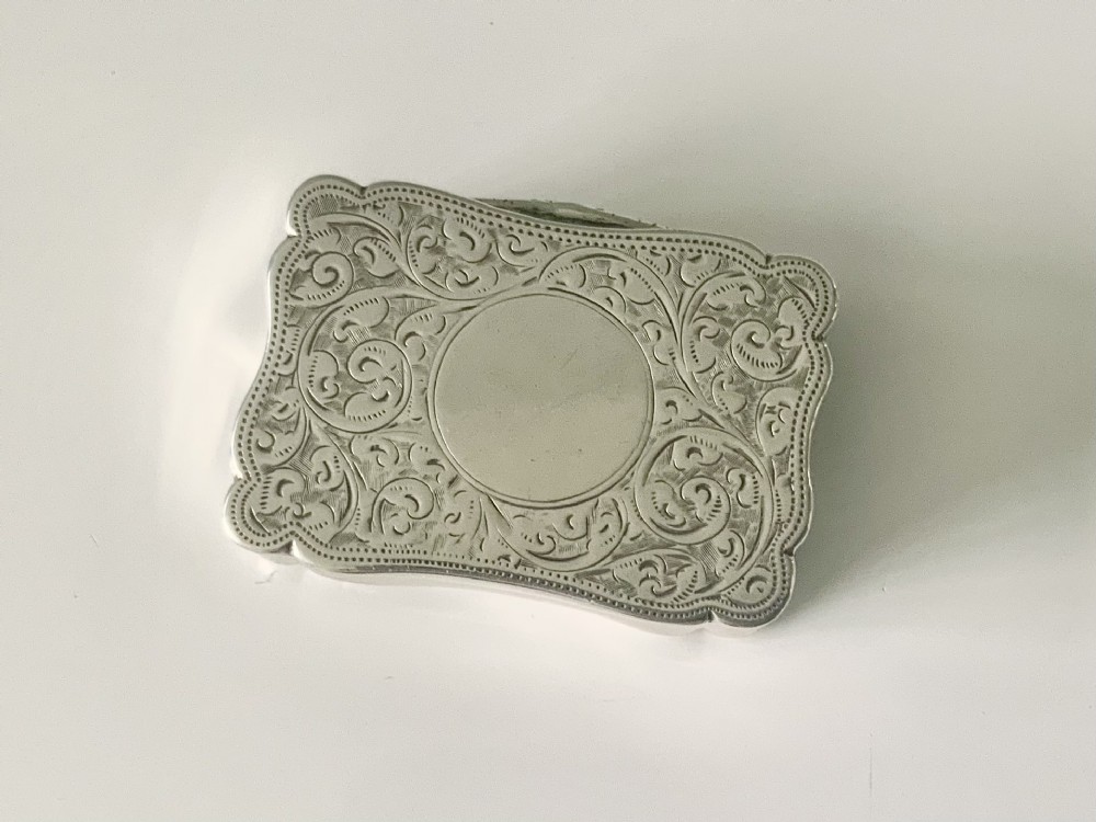 A rectangular sterling silver snuff box with a hinged lid, featuring detailed foliate engraving, a scalloped border, and a plain central circular cartouche.