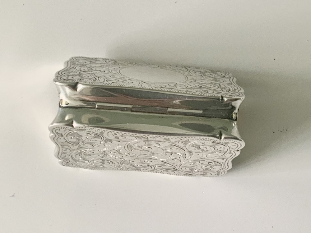 A rectangular sterling silver snuff box with a hinged lid, featuring detailed foliate engraving, a scalloped border, and a plain central circular cartouche. View VI