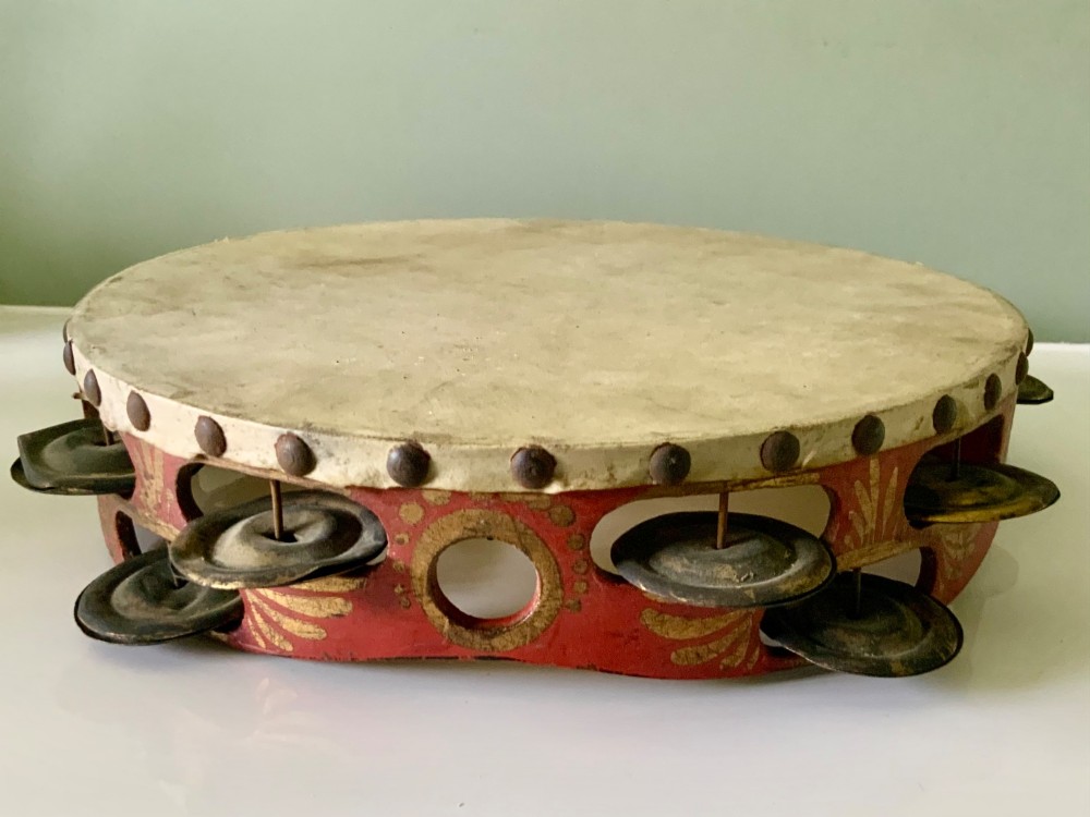 An antique Regency circular wooden tambourine featuring hand-painted red and gold floral patterns, metal jingles set into the rim, and a taut vellum head secured with brass studs. View V