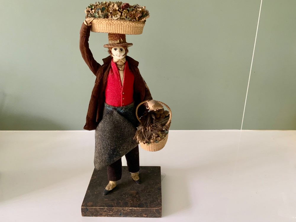 A 19th-century Neapolitan creche figure representing a street vendor dressed in fabric garments, holding woven straw baskets overhead and at his side, mounted on a square base.