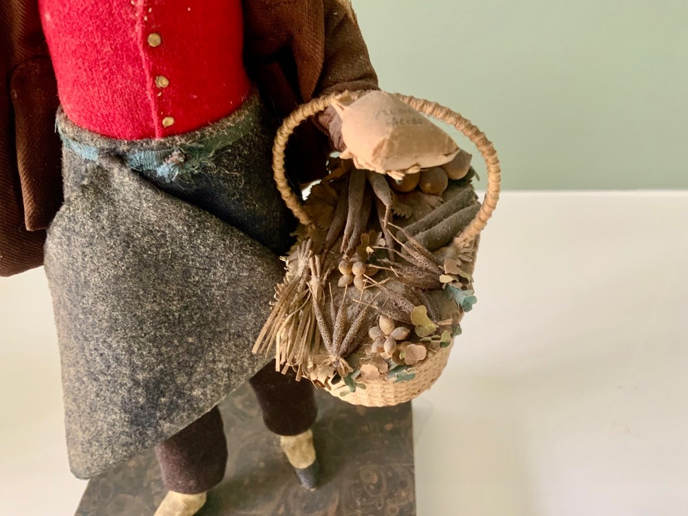 A 19th-century Neapolitan creche figure representing a street vendor dressed in fabric garments, holding woven straw baskets overhead and at his side, mounted on a square base. View II