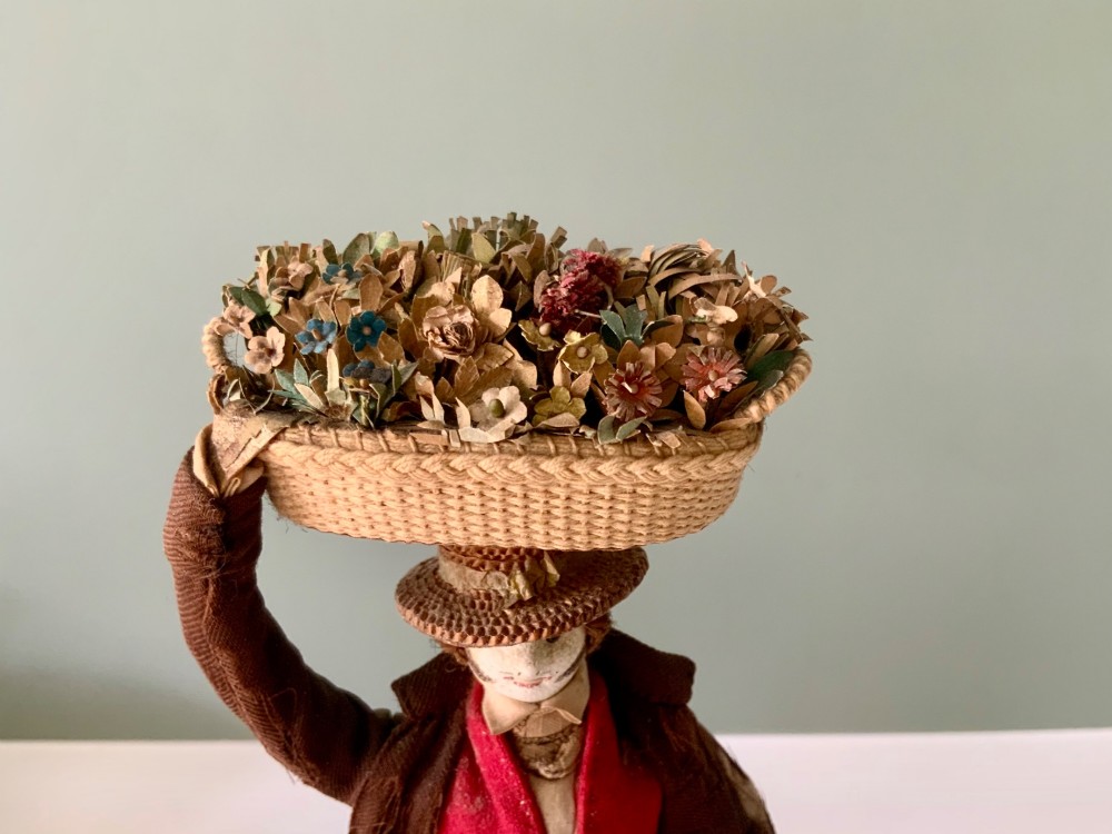 A 19th-century Neapolitan creche figure representing a street vendor dressed in fabric garments, holding woven straw baskets overhead and at his side, mounted on a square base. View III