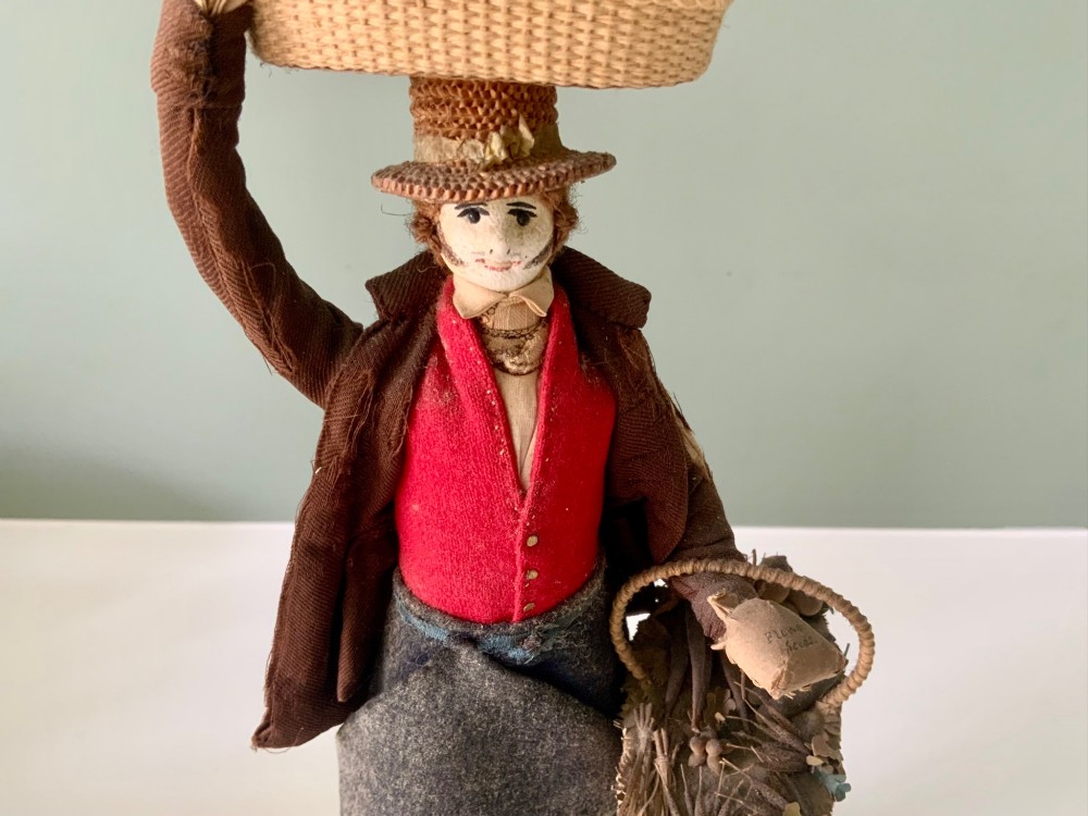 A 19th-century Neapolitan creche figure representing a street vendor dressed in fabric garments, holding woven straw baskets overhead and at his side, mounted on a square base. View IV