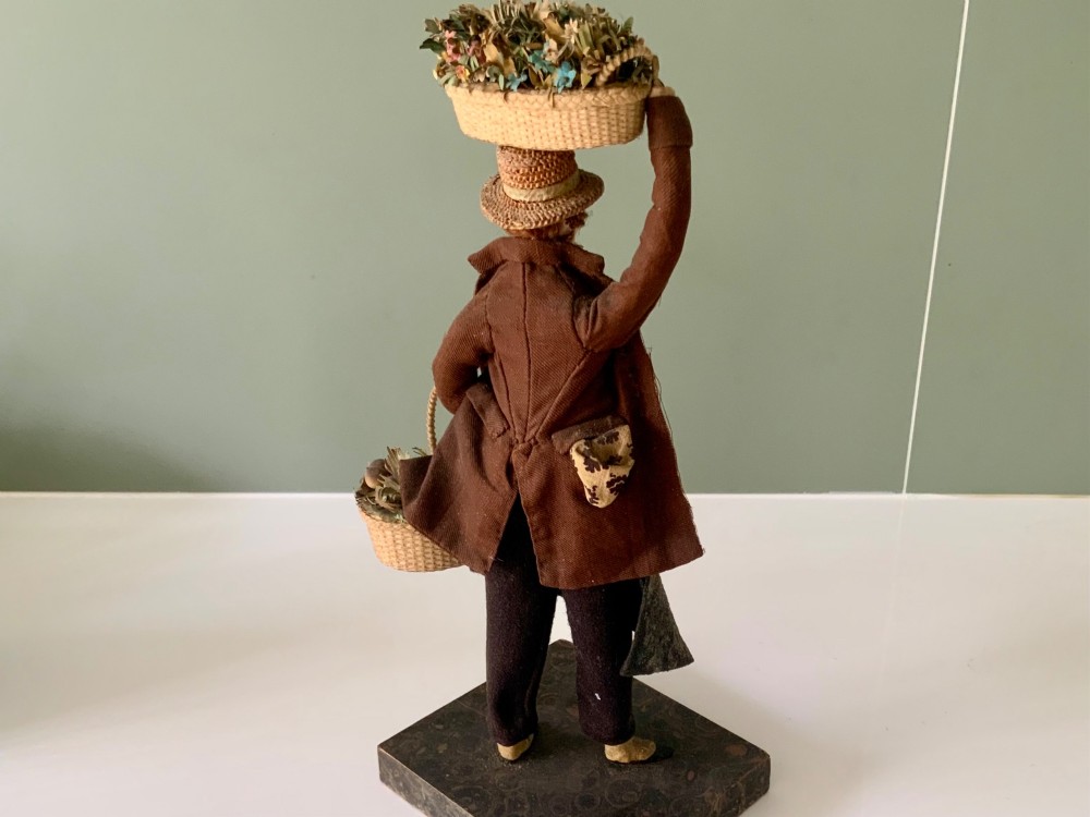 A 19th-century Neapolitan creche figure representing a street vendor dressed in fabric garments, holding woven straw baskets overhead and at his side, mounted on a square base. View V