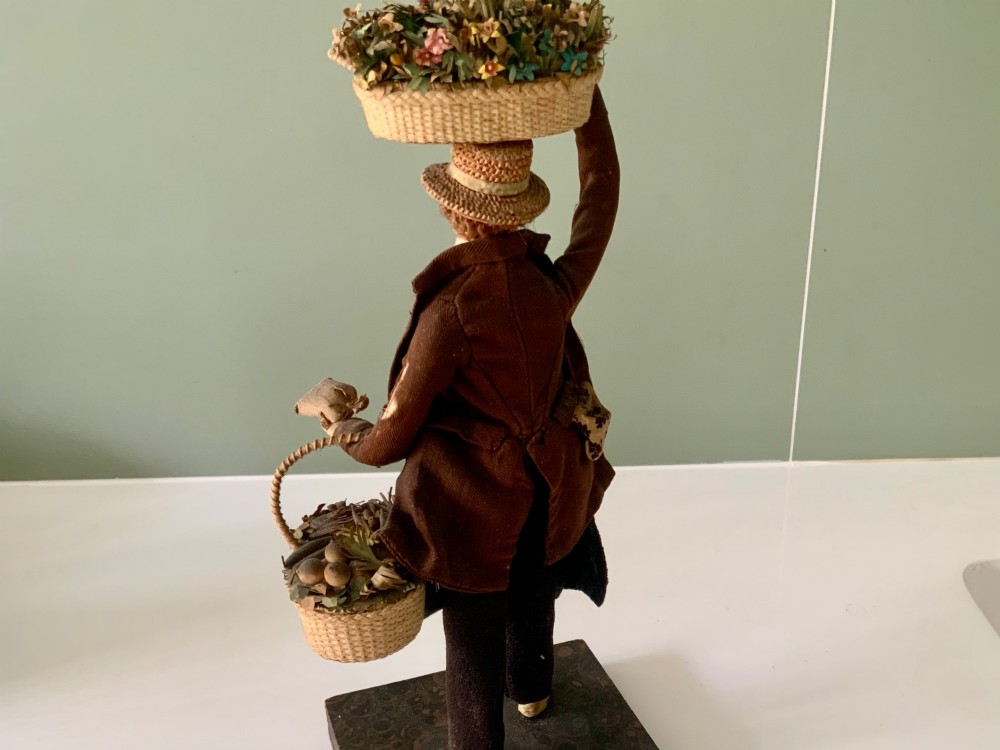 A 19th-century Neapolitan creche figure representing a street vendor dressed in fabric garments, holding woven straw baskets overhead and at his side, mounted on a square base. View VI