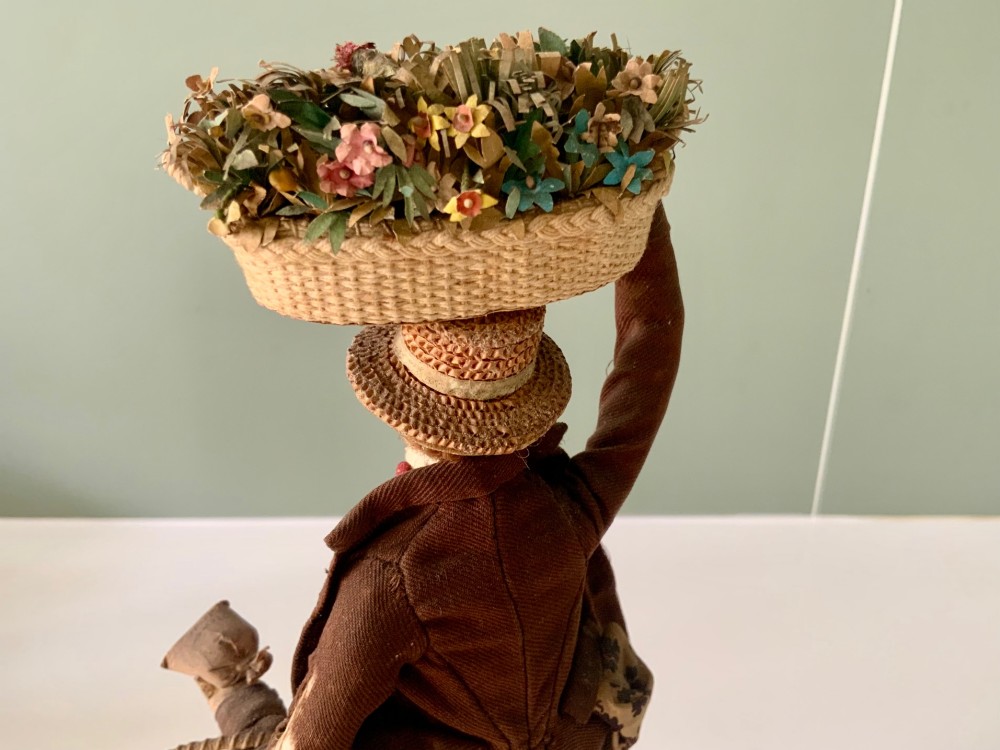 A 19th-century Neapolitan creche figure representing a street vendor dressed in fabric garments, holding woven straw baskets overhead and at his side, mounted on a square base. View VII
