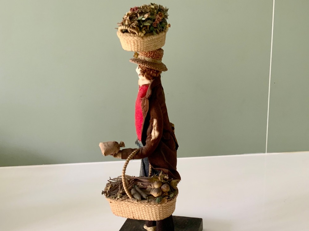 A 19th-century Neapolitan creche figure representing a street vendor dressed in fabric garments, holding woven straw baskets overhead and at his side, mounted on a square base. View VIII