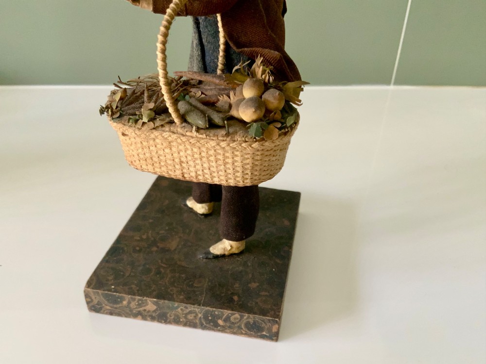A 19th-century Neapolitan creche figure representing a street vendor dressed in fabric garments, holding woven straw baskets overhead and at his side, mounted on a square base. View IX