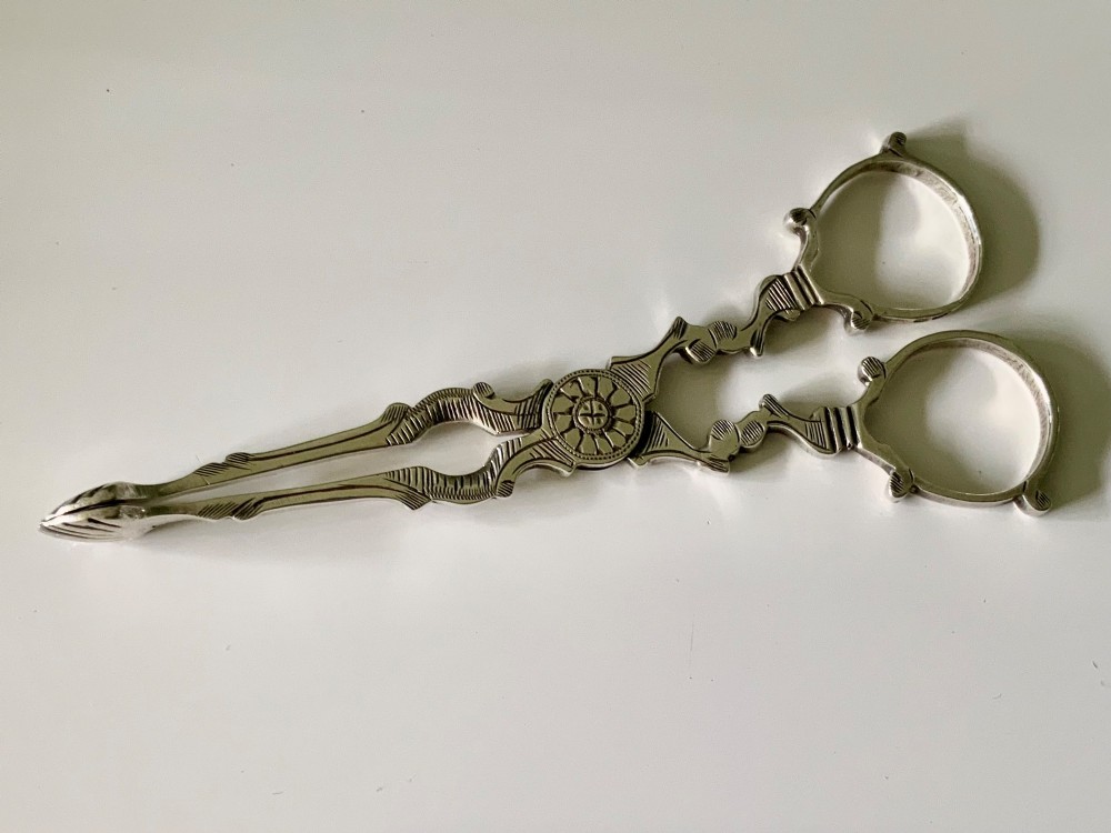 A pair of Georgian-style silver scissor-action sugar tongs with chased engraving and shell-shaped grips against a neutral background.