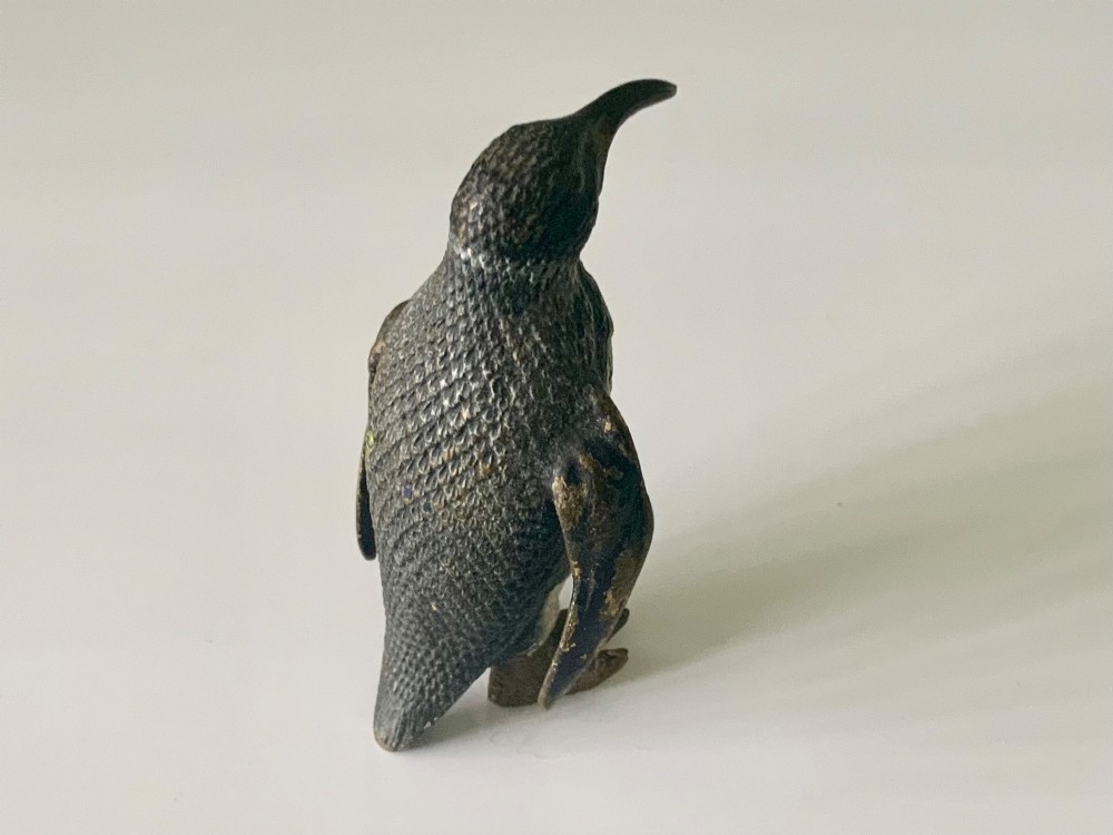 A small cold-painted bronze figurine depicting a standing penguin with textured feather details in dark brown, black, and off-white paint. View IV