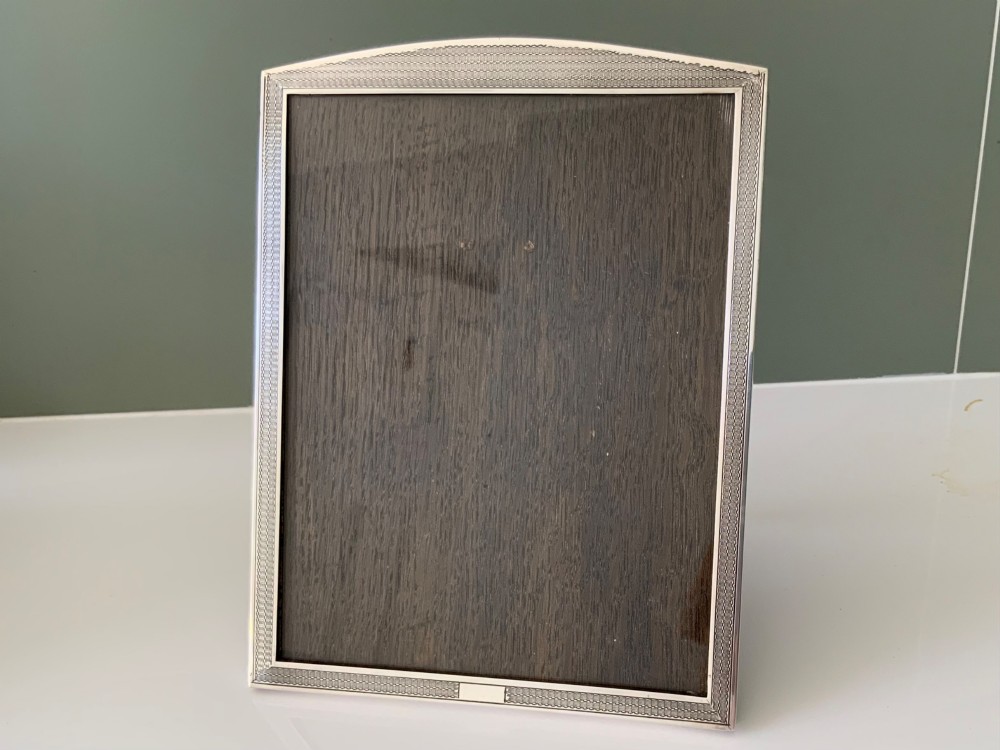 An Art Deco silver photo frame with a textured engine-turned border, an arched top profile, and a dark wooden interior backing.