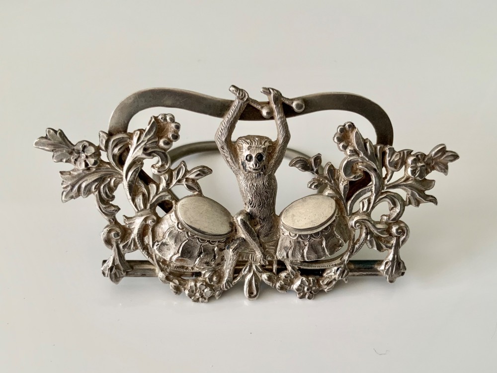 A cast silver menu holder featuring a central monkey figure playing drums framed by foliate scrollwork.