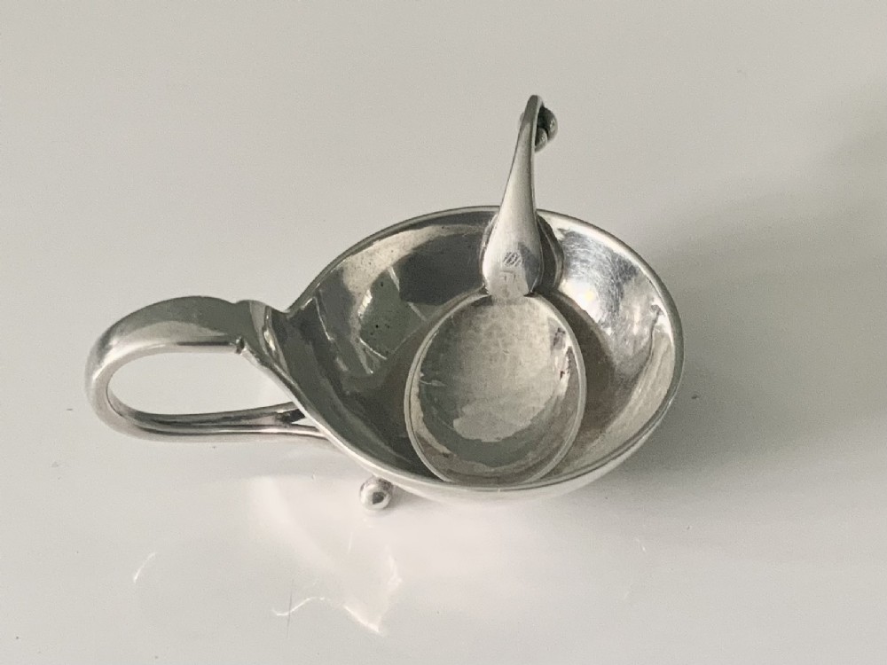 A Georg Jensen sterling silver open salt cellar with a curved handle and small spherical feet, containing a matching silver salt spoon.
