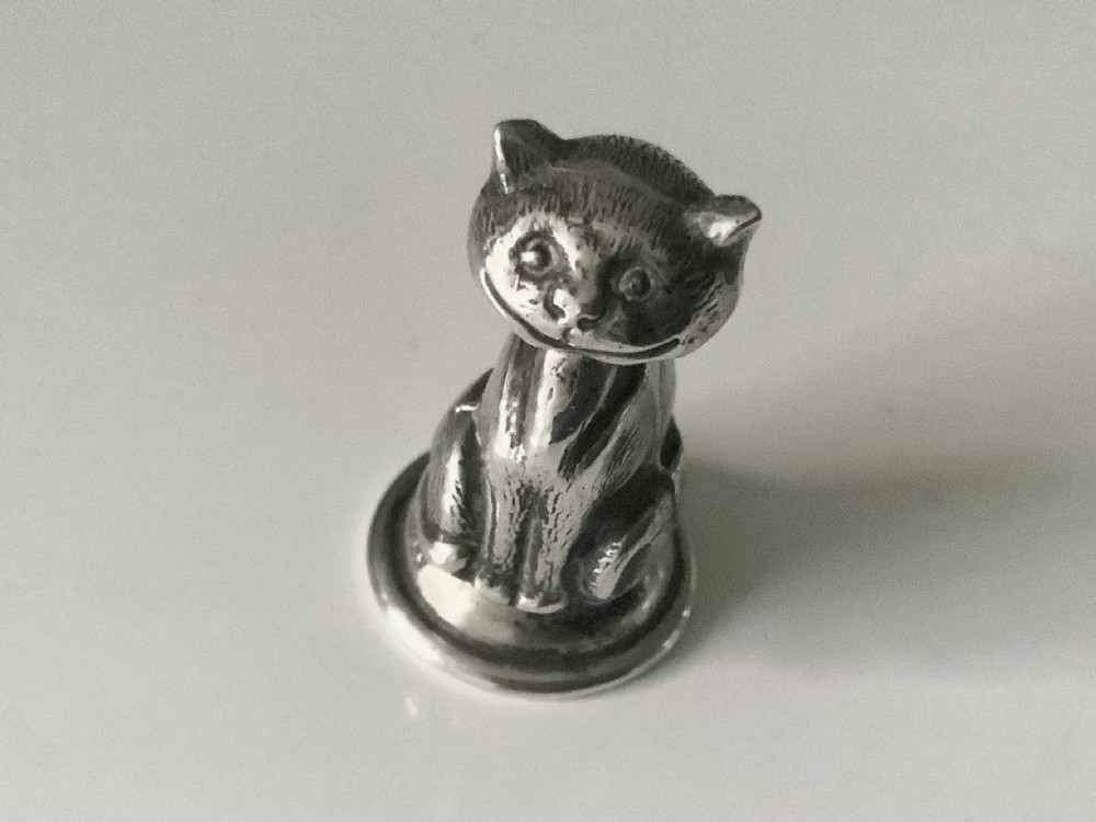 A small cast sterling silver figurine of a smiling Cheshire cat seated on a rounded wax seal base. View II