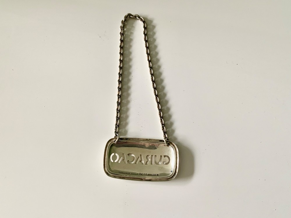 A rectangular sterling silver decanter label with cut-out lettering reading CURACAO suspended by a fine silver chain. View II