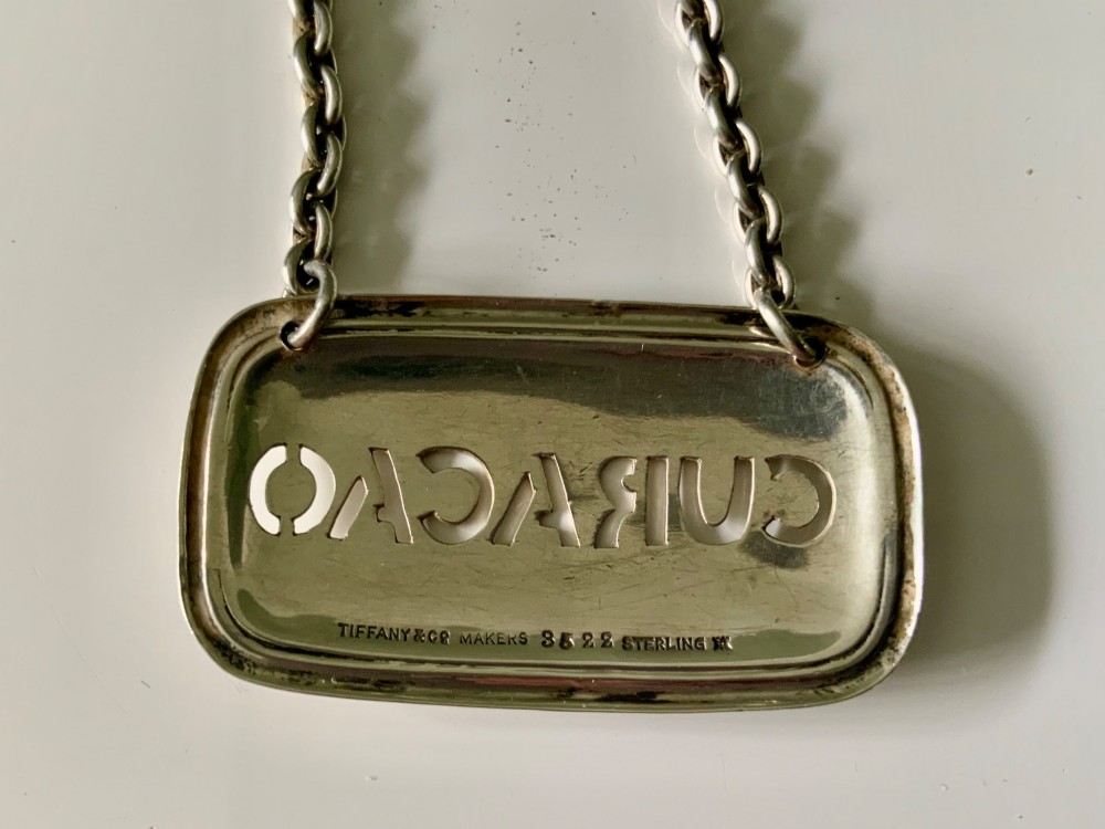 A rectangular sterling silver decanter label with cut-out lettering reading CURACAO suspended by a fine silver chain. View III