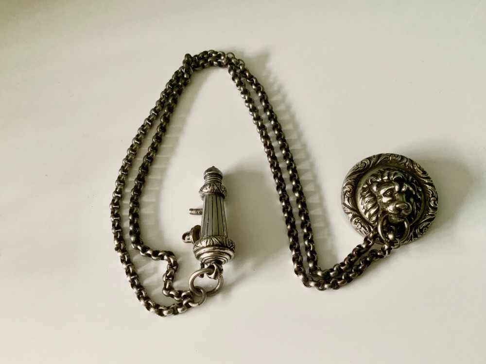 A silver-plated military whistle with fluted details connected by a metal link chain to a circular lion mask boss mount.
