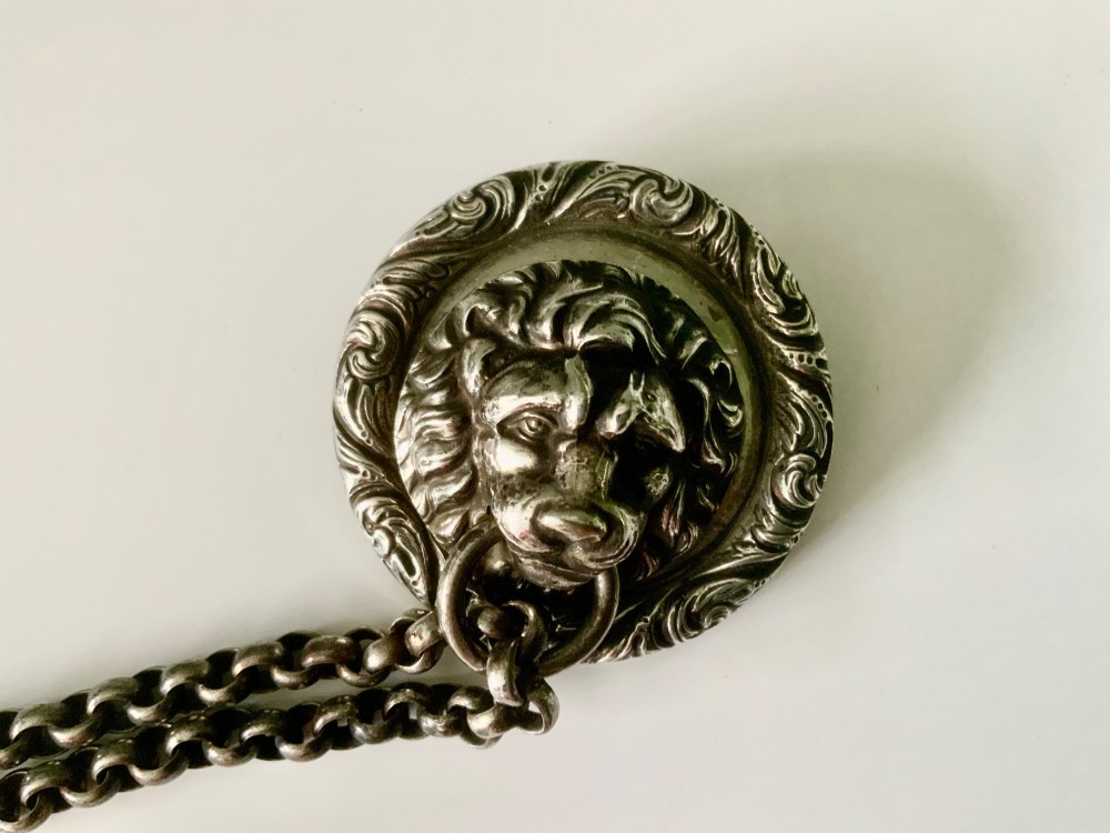 A silver-plated military whistle with fluted details connected by a metal link chain to a circular lion mask boss mount. View II