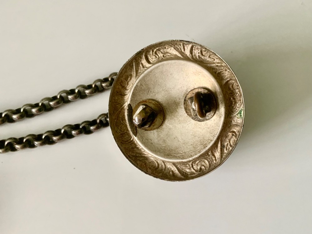 A silver-plated military whistle with fluted details connected by a metal link chain to a circular lion mask boss mount. View III