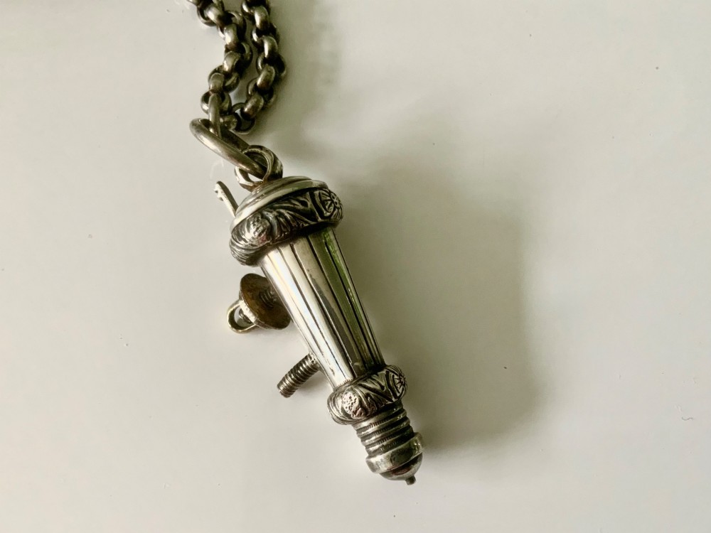 A silver-plated military whistle with fluted details connected by a metal link chain to a circular lion mask boss mount. View V