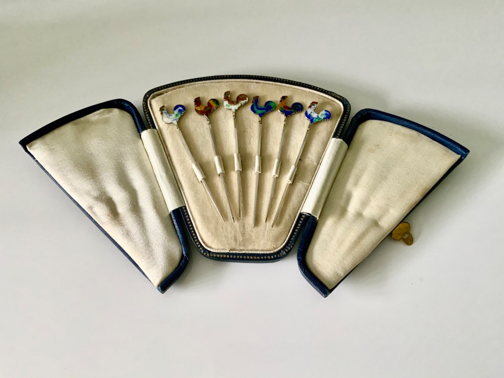 A set of six silver cocktail sticks featuring colourful enamelled cockerel finials, displayed inside an open fitted fan-shaped blue leather case.