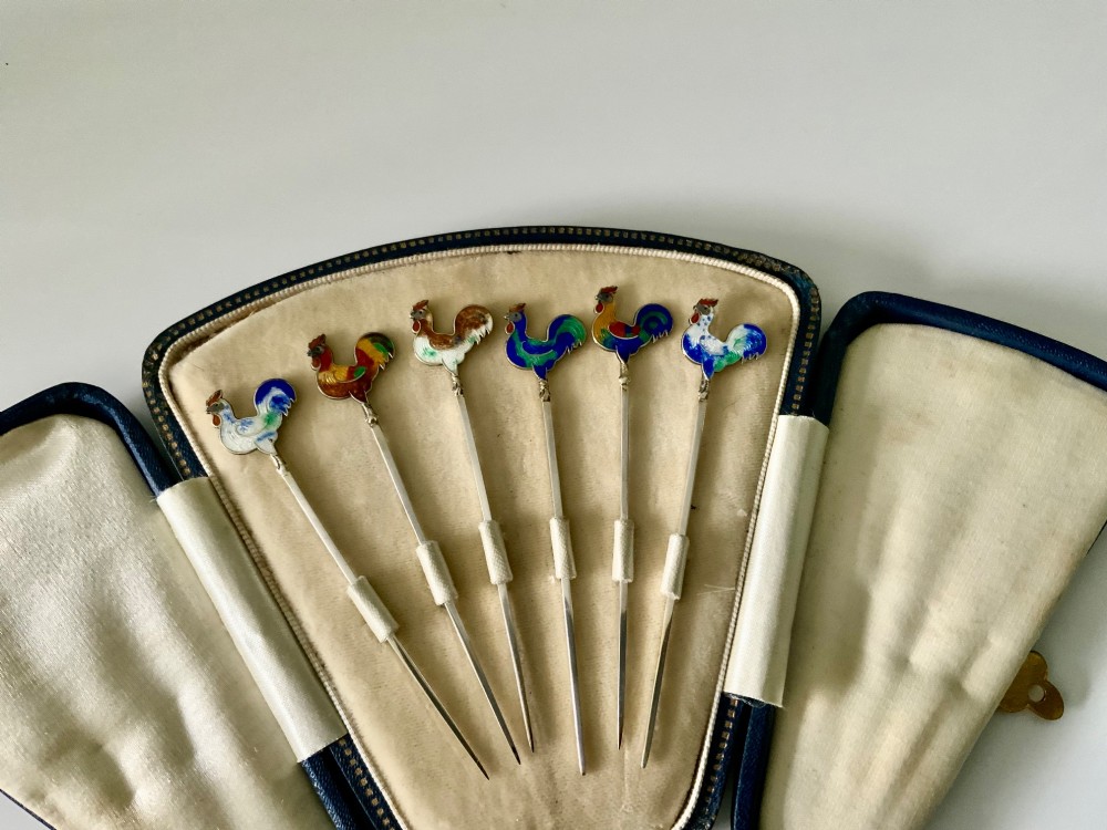 A set of six silver cocktail sticks featuring colourful enamelled cockerel finials, displayed inside an open fitted fan-shaped blue leather case. View II