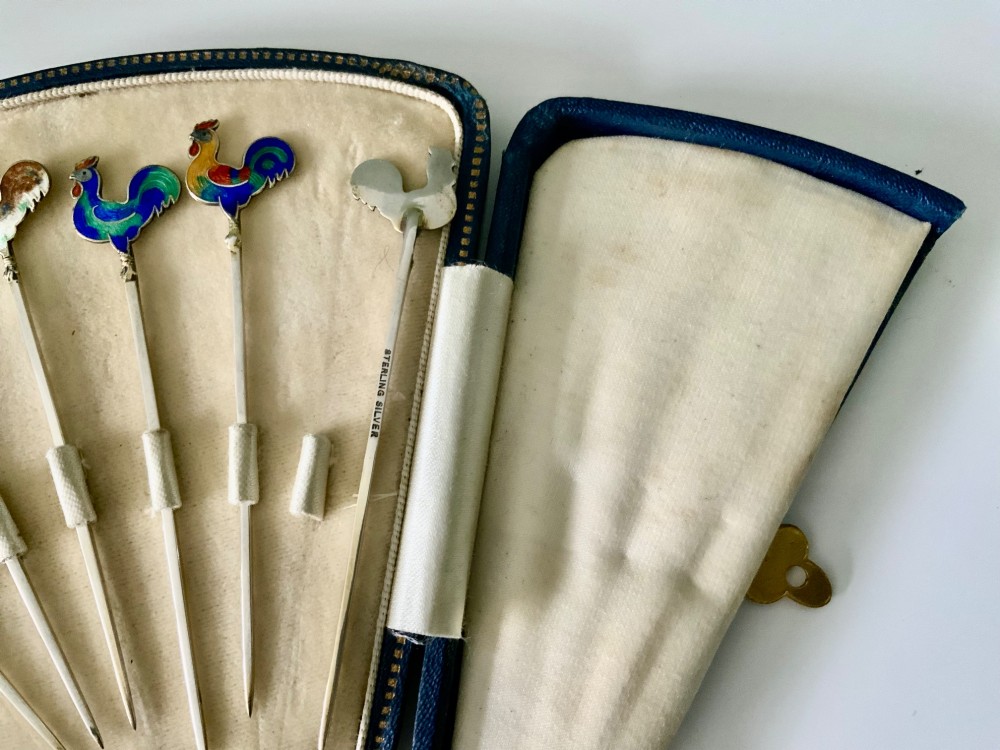 A set of six silver cocktail sticks featuring colourful enamelled cockerel finials, displayed inside an open fitted fan-shaped blue leather case. View III
