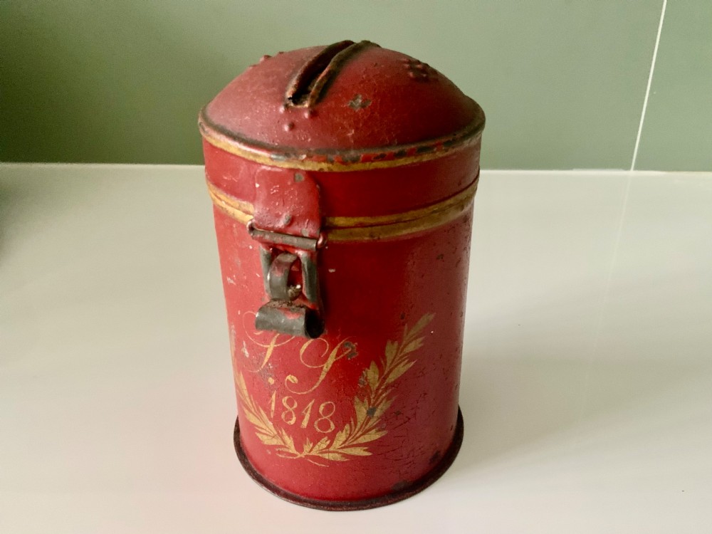 A cylindrical red painted toleware money box dated 1818 featuring a domed top with coin slot, metal latch, and gilded leaf decoration.