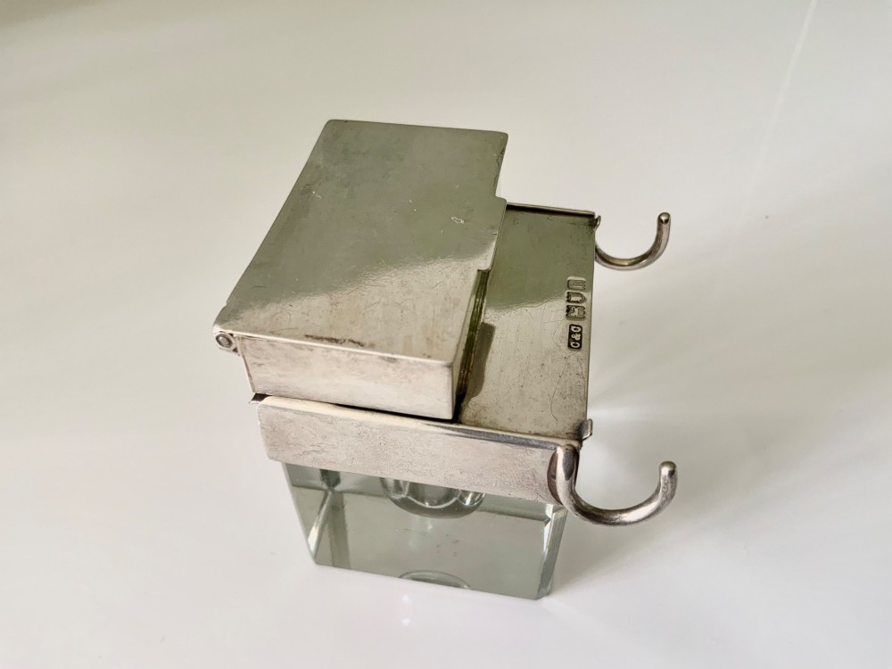 A square clear glass inkwell featuring a hallmarked sterling silver hinged lid, integrated stamp compartment, and side pen rests. View V