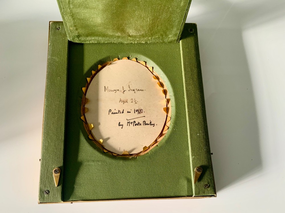 An oval framed portrait miniature depicting a fair-haired child wearing a white shirt and blue bow tie, presented within a cream fabric mount and a carved gilded rectangular frame. View II