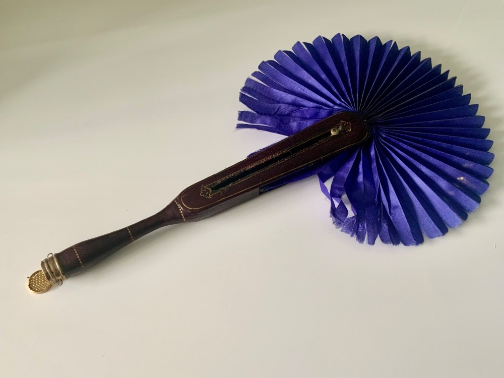 A 19th-century French hand fan featuring a dark brown leather handle with an integrated gilt metal vinaigrette grille at the base and a pleated purple leaf.