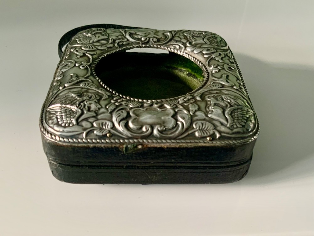 An embossed silver pocket watch stand featuring the Reynolds Angels cherub motif with a circular opening and velvet lining. View VIII