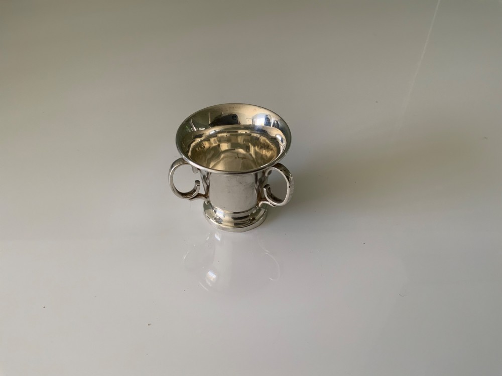 A polished miniature silver tyg featuring a flared rim, three looped handles, and a stepped pedestal base.