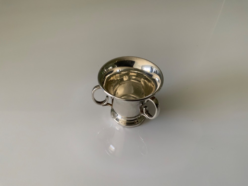 A polished miniature silver tyg featuring a flared rim, three looped handles, and a stepped pedestal base. View II
