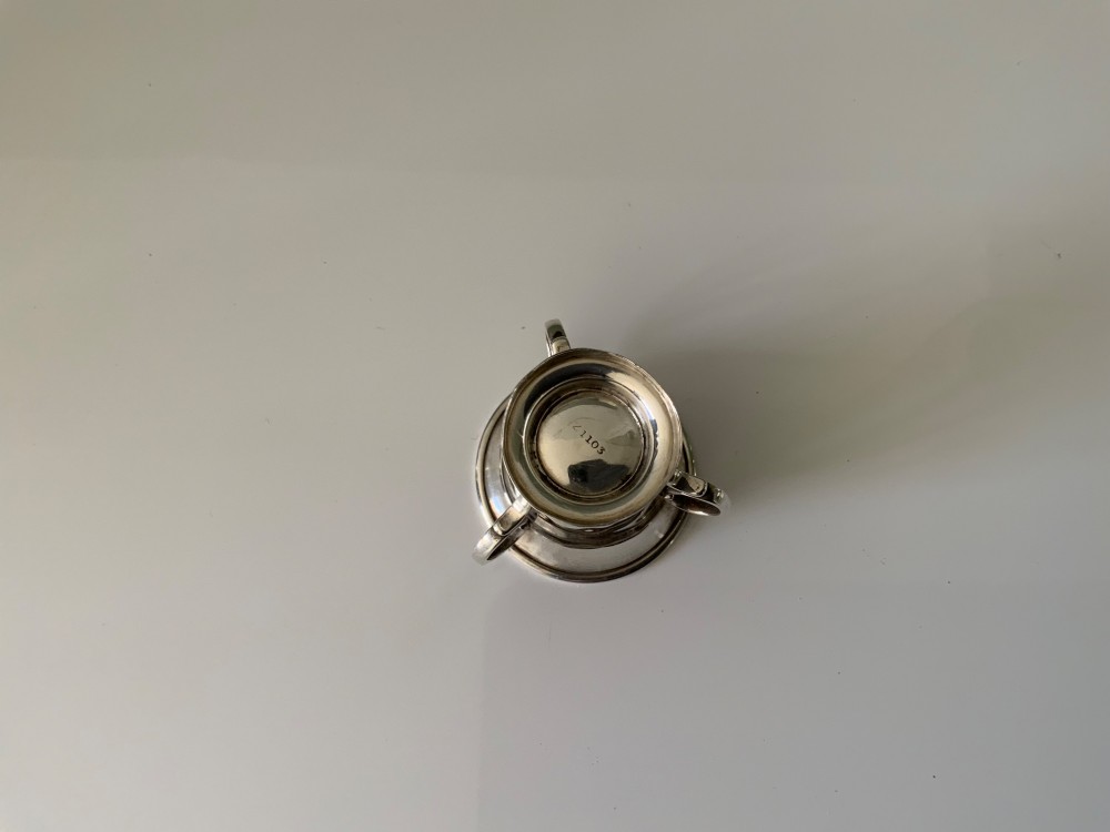 A polished miniature silver tyg featuring a flared rim, three looped handles, and a stepped pedestal base. View V