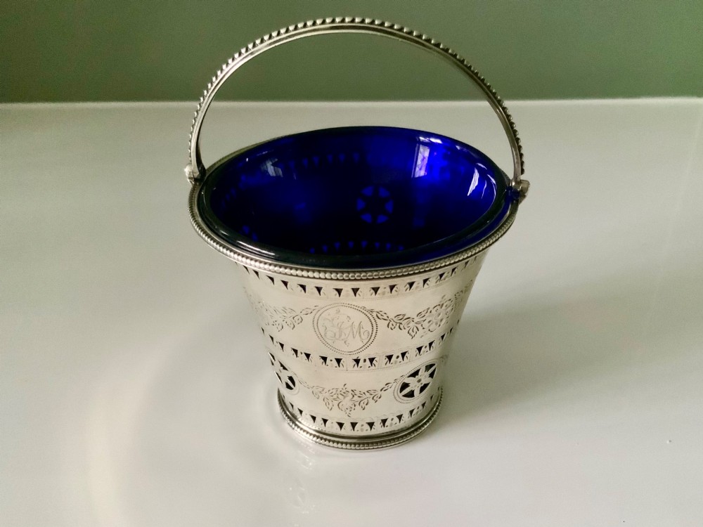 A Georgian sterling silver pierced cream pail featuring bright-cut engraved decoration, an overhead beaded handle, and a removable cobalt blue glass liner. View II