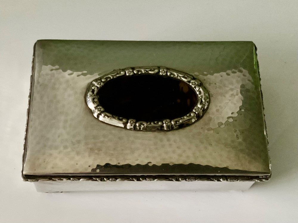 A rectangular silver plated box featuring a hand-hammered lid and a dark oval central inset framed by decorative detailing.