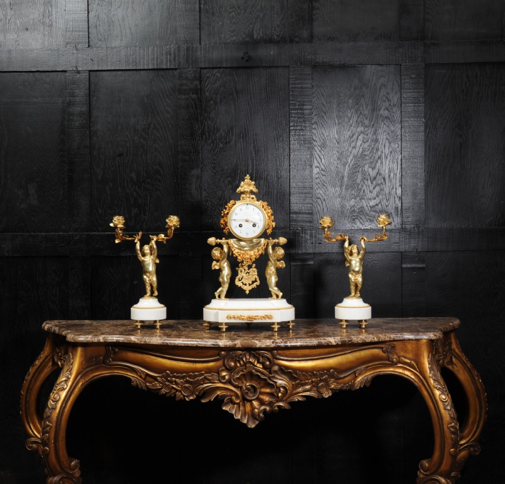 An antique French gilt bronze and white marble clock garniture set featuring a central mantel clock flanked by two matching cherub candelabra. View II