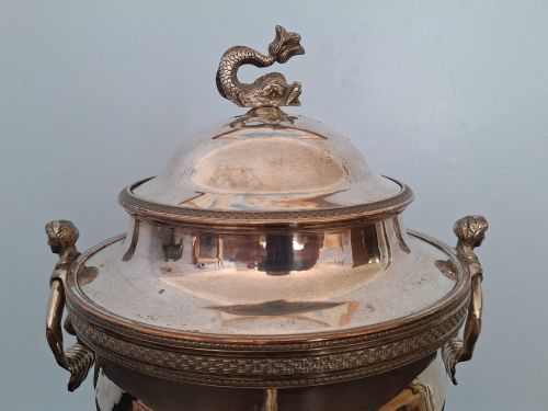 fig 2. An antique silver-plated Regency tea urn featuring a top dolphin finial, classical figure side handles, a front spigot, and a square base supported by ball feet.