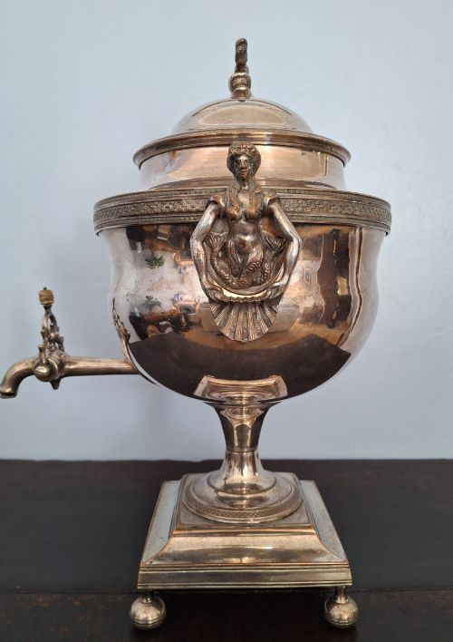 fig 3. An antique silver-plated Regency tea urn featuring a top dolphin finial, classical figure side handles, a front spigot, and a square base supported by ball feet.