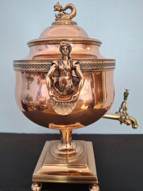 fig 2. An antique polished copper and brass tea urn featuring a urn-shaped body, crest finial, side handles, and a spigot, resting on a square pedestal base.