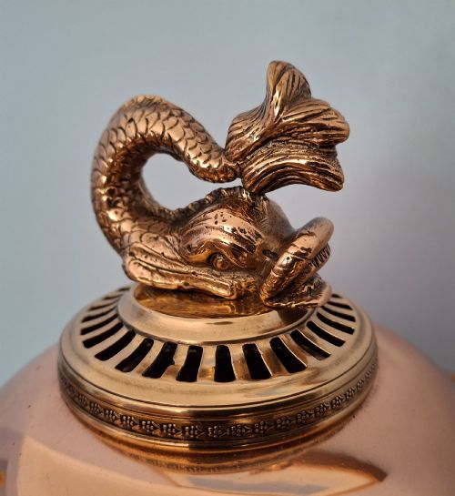 fig 4. An antique polished copper and brass tea urn featuring a urn-shaped body, crest finial, side handles, and a spigot, resting on a square pedestal base.