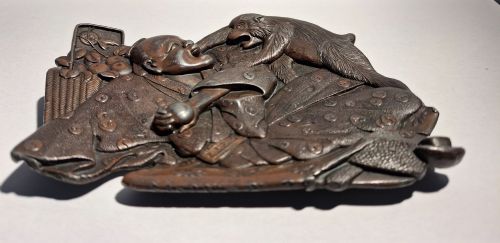 fig 1. A dark bronze Japanese Takazogan metal tray depicting a figure in patterned robes reacting to a monkey perched on his shoulder.