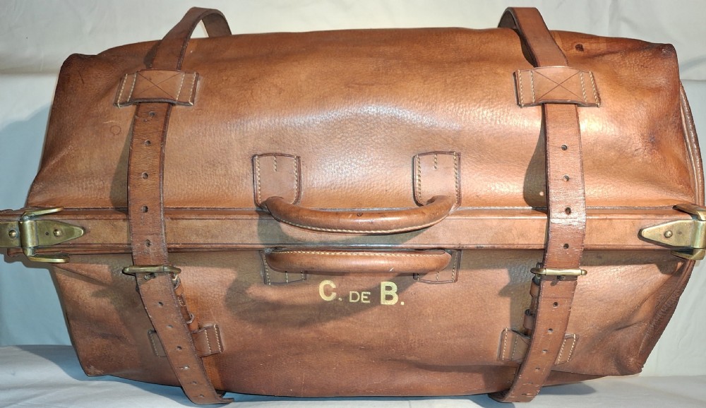 A warm brown leather Gladstone bag featuring twin buckle straps, a brass central lock mechanism, a thick handle, and gold initials. View II
