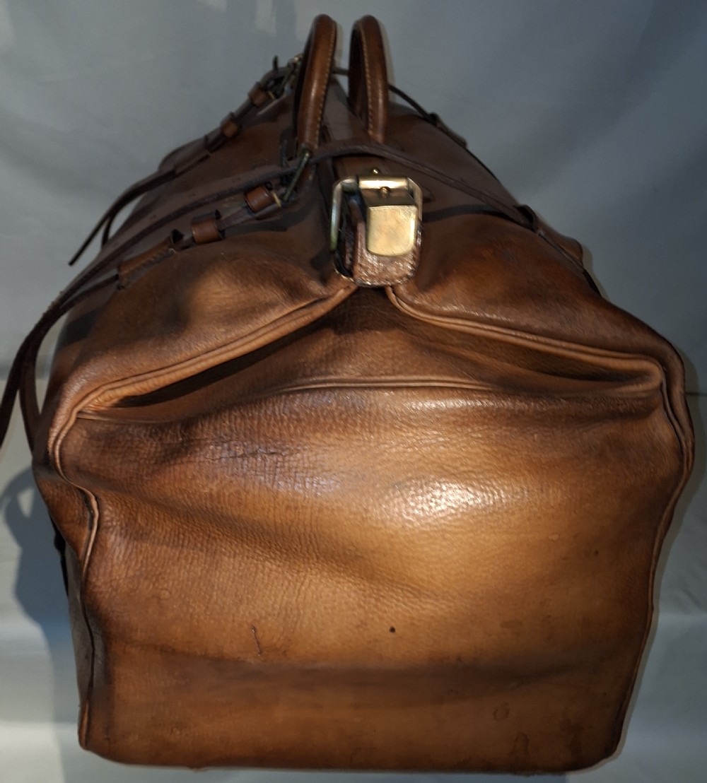 A warm brown leather Gladstone bag featuring twin buckle straps, a brass central lock mechanism, a thick handle, and gold initials. View III